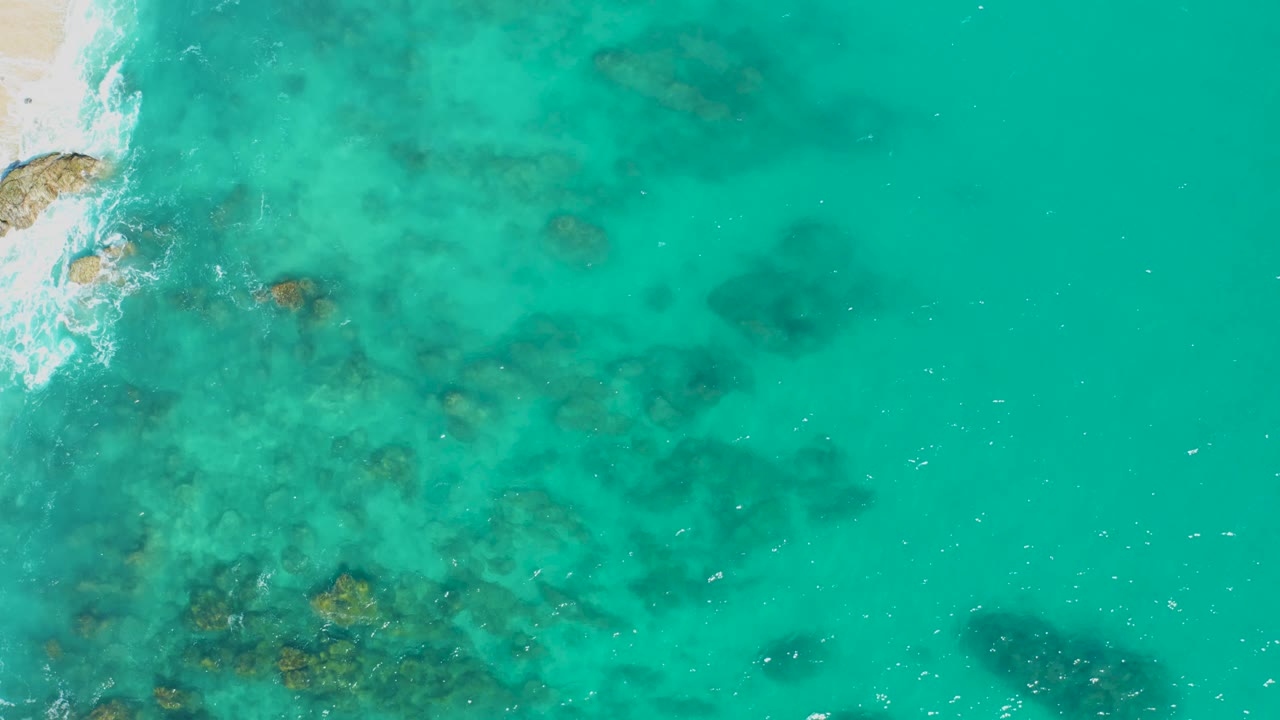 Aerial view in East Cape, Baja California Sur with open turquoise water dominating the frame, reef shadows below the surface, surf along the upper-left shore, and no visible people.