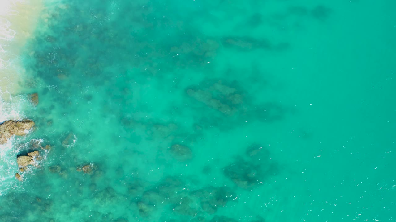 Overhead view in East Cape BCS Mexico with foamy water around left-side rocks, visible underwater formations in the center, open turquoise water to the right, and no visible people.