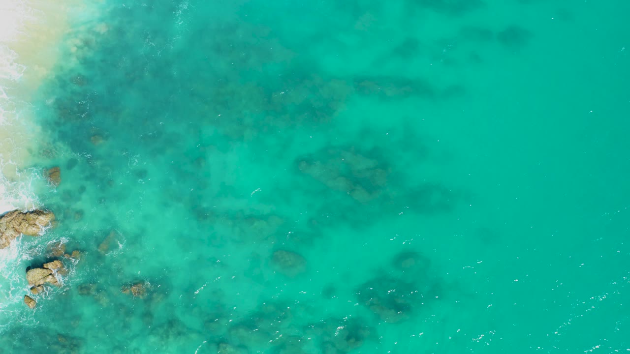 Overhead East Cape BCS Mexico shoreline with pale sand and foam at the left border, scattered darker underwater rocks through the middle, and no visible people.