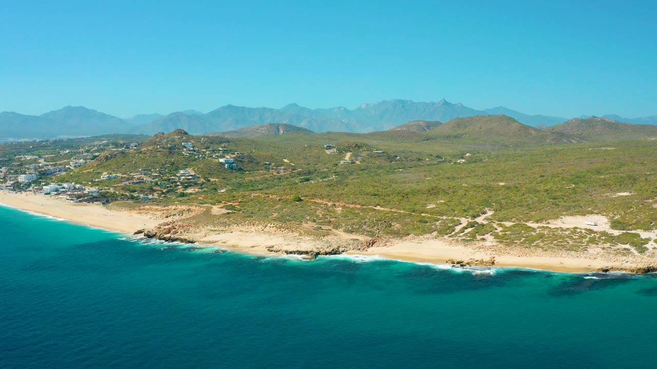 Aerial frame from East Cape BCS Mexico showing turquoise ocean below a sandy and rocky shoreline, green coastal hills, distant mountains, clear sky, and no visible people.