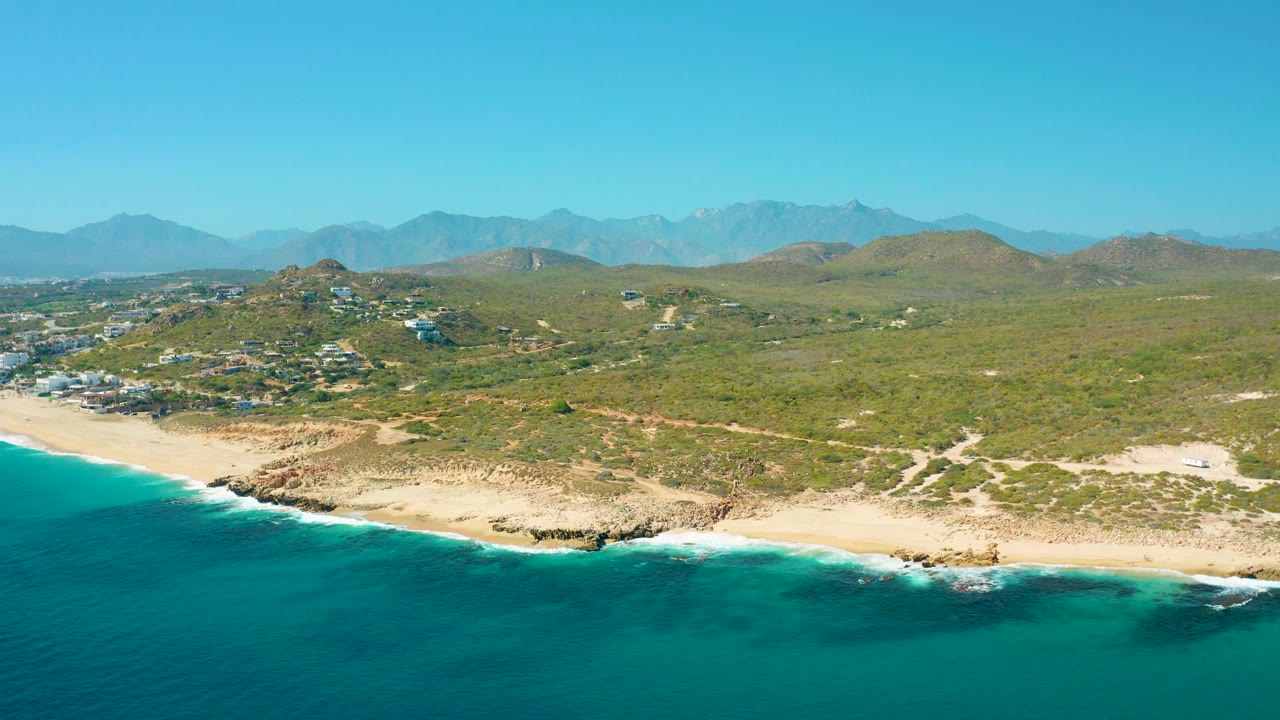 Opening aerial frame over East Cape BCS Mexico with turquoise water in the foreground, pale beach below green hills, distant mountains, clear sky, and no visible people.