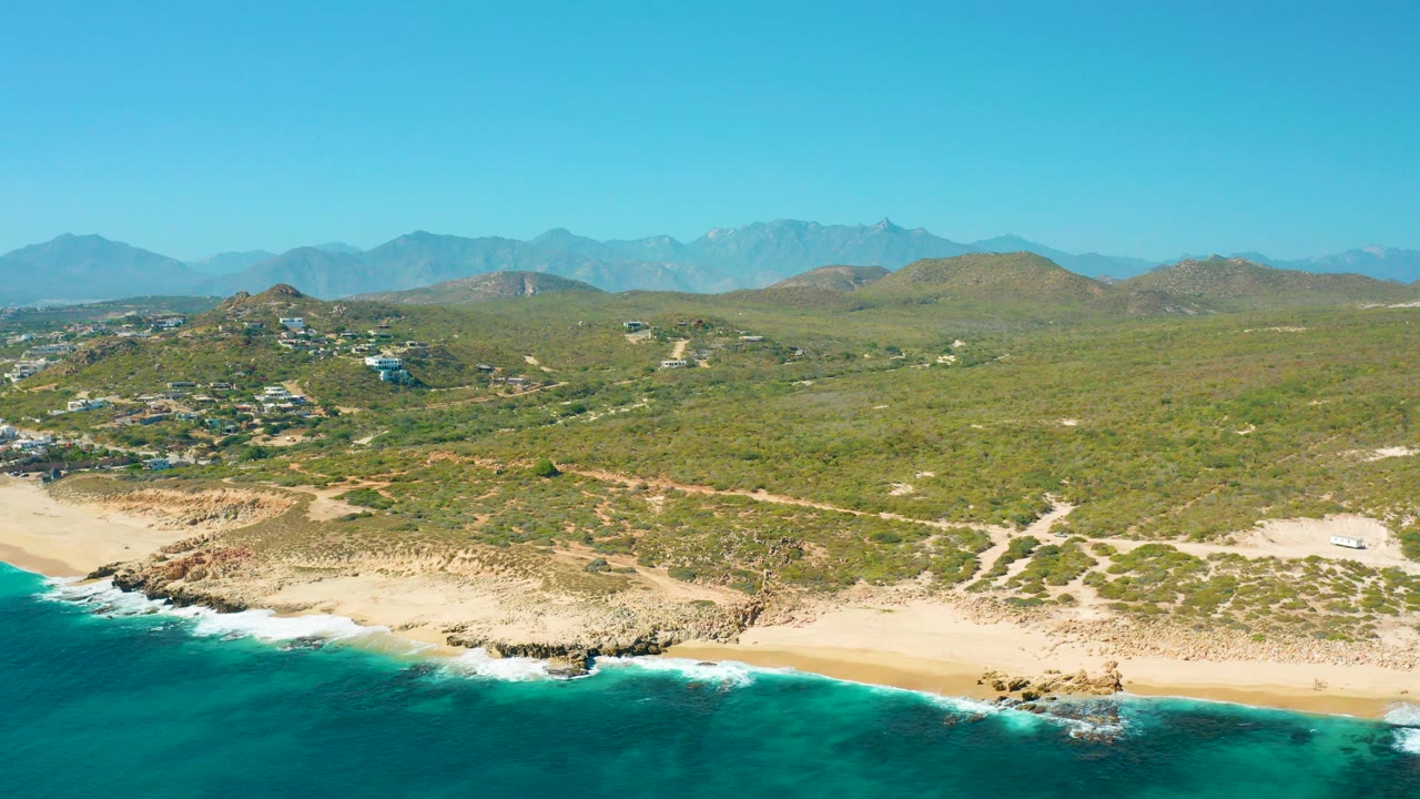 Aerial frame from East Cape, Baja California Sur, showing turquoise surf at the lower edge, a sandy beach and rocky shoreline, green hills inland, distant mountains, clear blue sky, and no visible people.