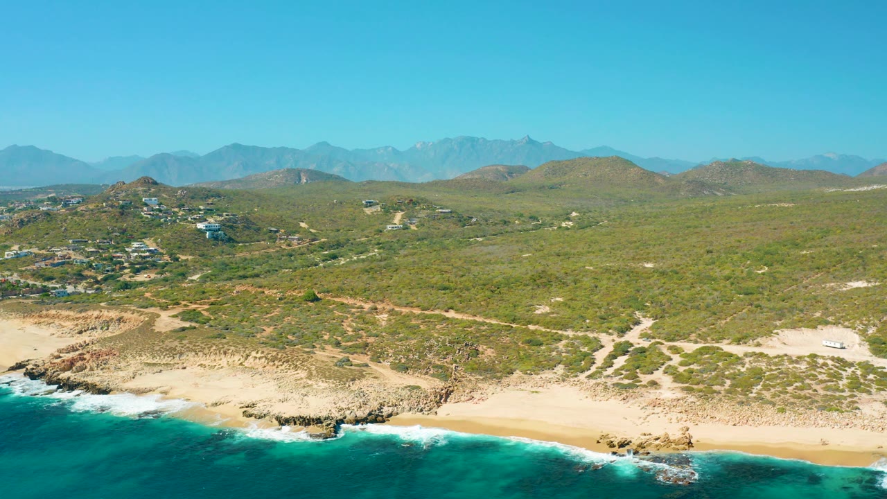 Drone view over ocean surf, beach, rocky ledges, hills, homes, and mountains.