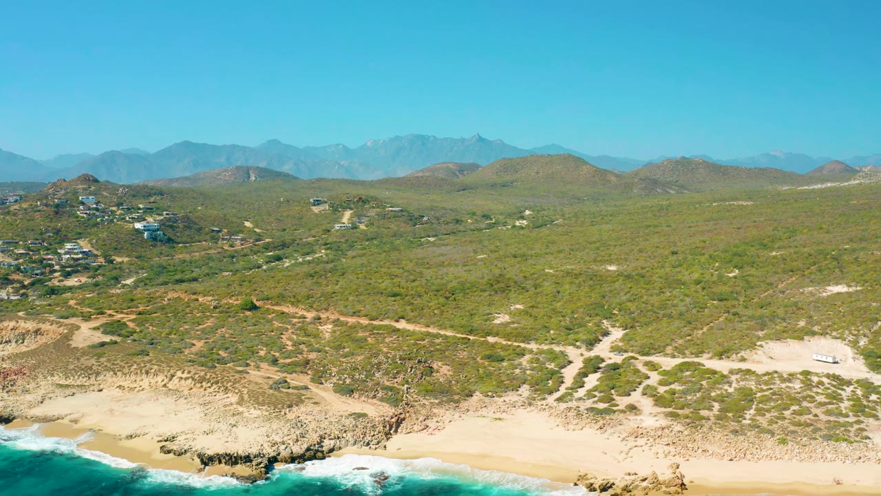 Drone view over sandy shore, rocky bluff, scrub hills, and mountain horizon
