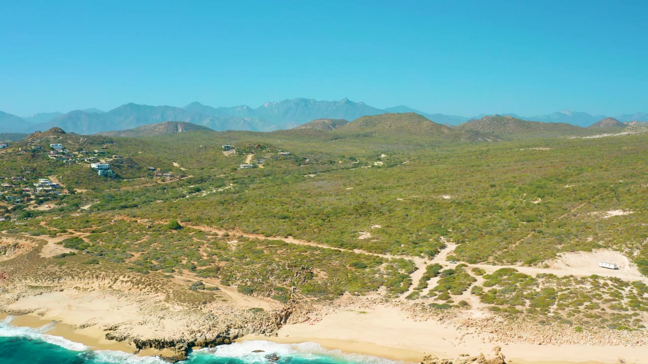 East Cape aerial frame with beach and surf still visible at the bottom, rocky tan shoreline, dirt paths through green desert vegetation, scattered structures to the left, hazy mountains, and no visible people.