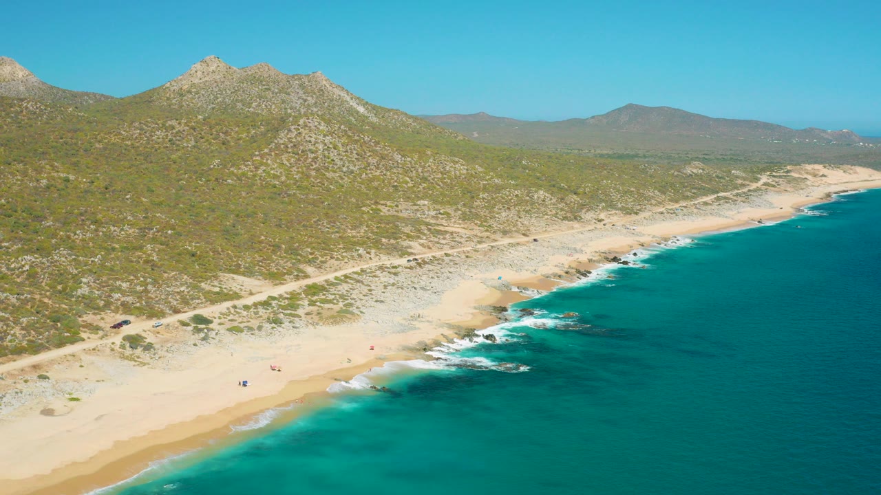Aerial view over East Cape, Baja California Sur, Mexico with turquoise water on the right, a pale sandy beach below, tiny beachgoers near the surf, and scrubby hills behind.