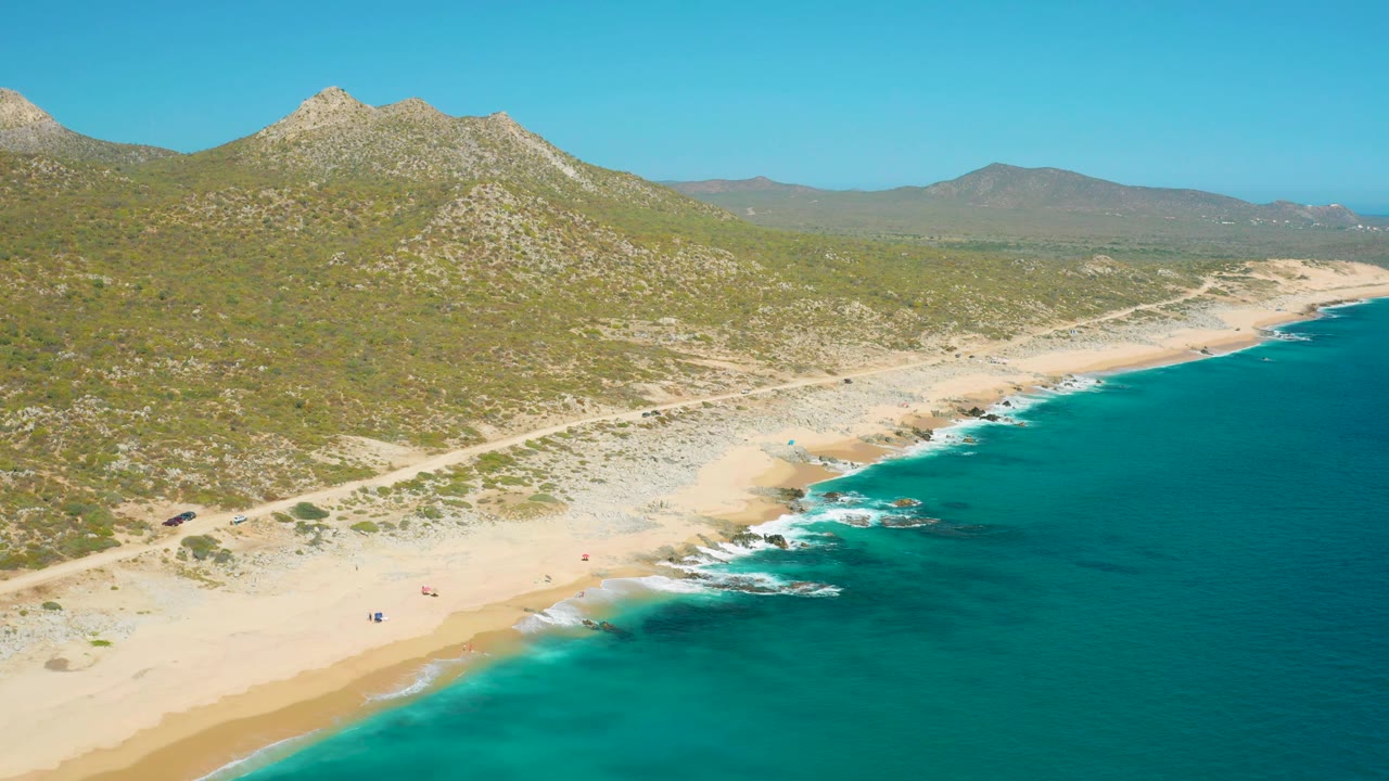 East Cape aerial view with the ocean occupying the right half, a thin surf line against the sand, tiny beachgoers below, and low mountains stretching across the horizon.