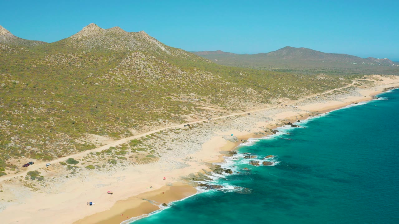 Aerial frame of East Cape BCS Mexico showing a sandy beach curving below green hills, turquoise water on the right, and small beachgoers and umbrellas on the sand.