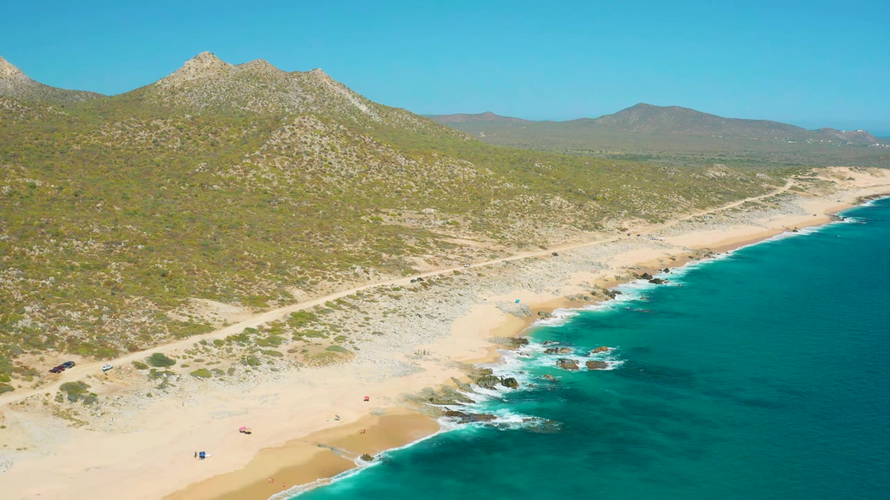 Wide drone view of beach, surf, dirt road, and mountains.