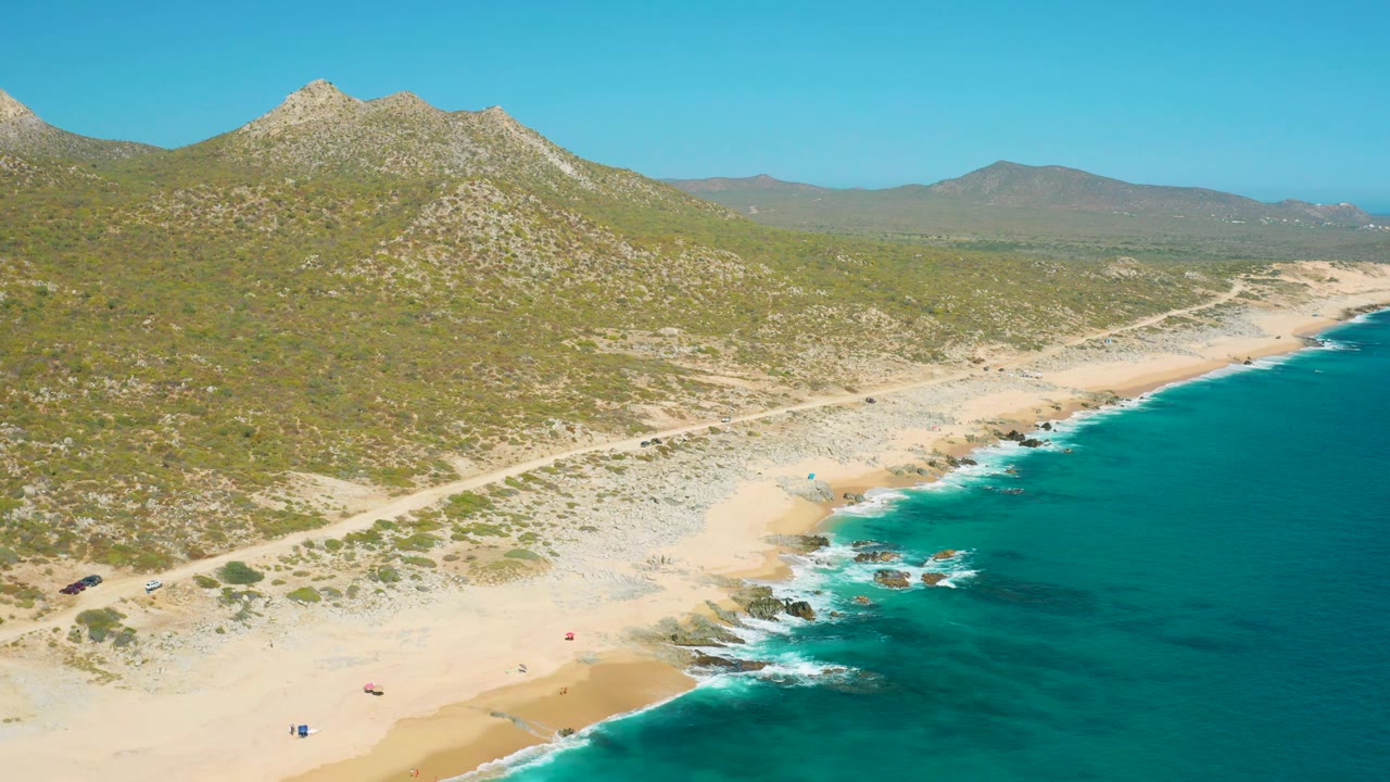 East Cape BCS Mexico coastline from above, with deeper blue water filling the right side, white wash at the rocks, scattered umbrellas on the beach, and mountains spanning the horizon.