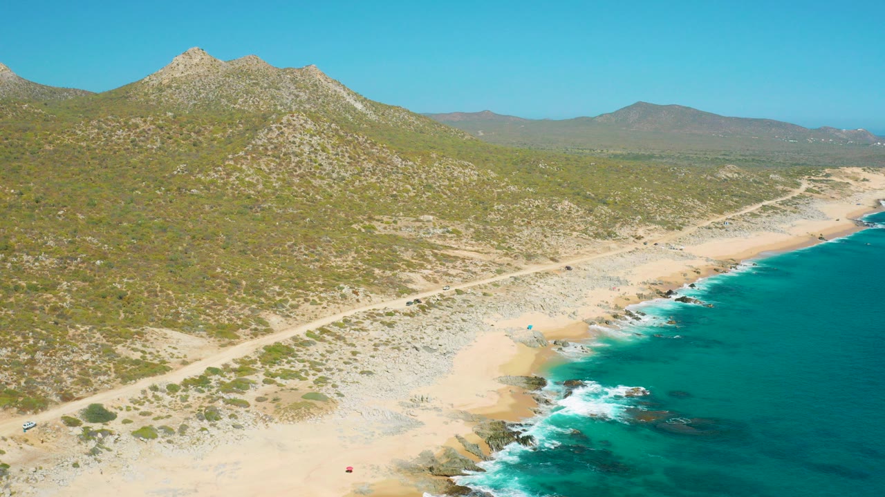 Aerial view of East Cape, Baja California Sur with a dirt road crossing scrubland above a sandy beach, turquoise surf on the right, mountains in the background, and tiny beachgoers visible.