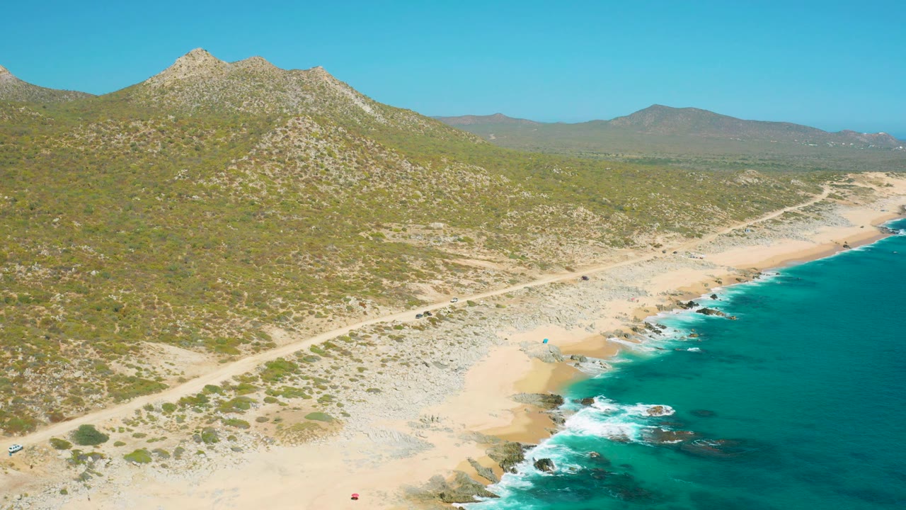 Aerial East Cape view with the ocean filling the right edge, waves breaking along sand and rocks, the dirt road running diagonally, scrubland rising toward rocky mountains, and tiny people visible near the beach.