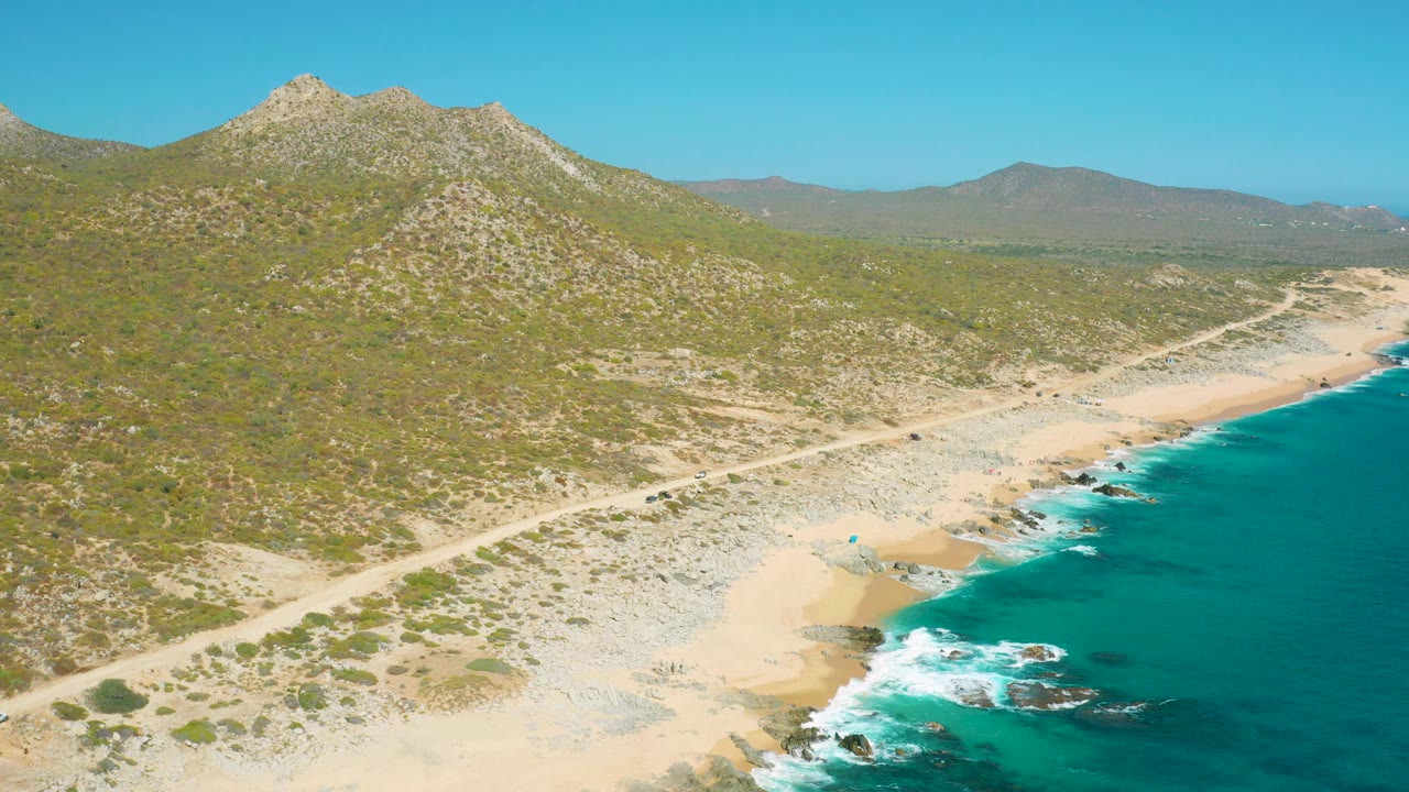 High coastal view with a dirt road, sandy shore, surf, and mountains.