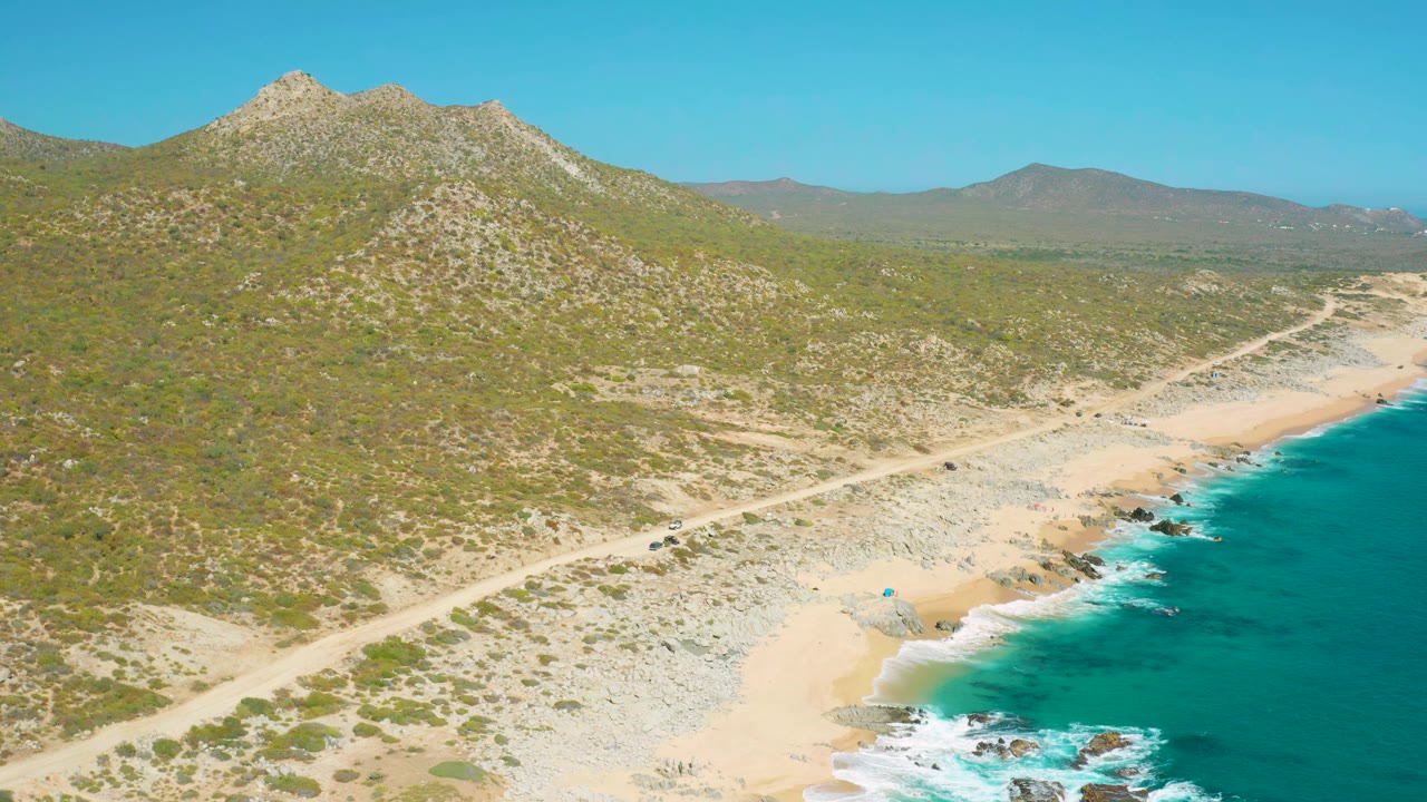 At 0:01, the East Cape BCS Mexico shoreline remains in a high aerial frame, with foamy waves edging the beach, the road crossing the midground, hills behind it, and no visible people.
