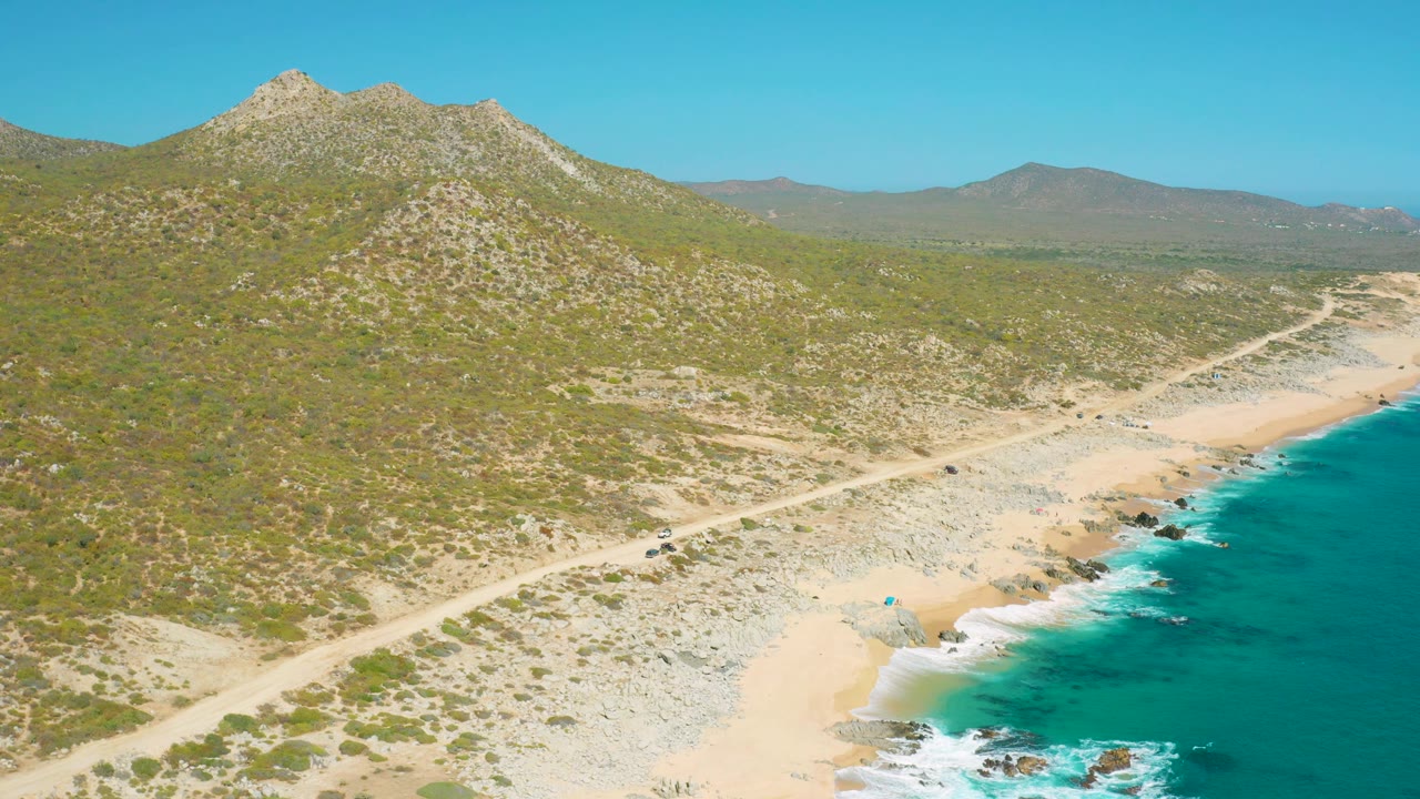 At 0:02, the aerial view shows the beach and road slightly farther along the coast, with blue-green water at frame right, rocky hillside above, and no visible people.