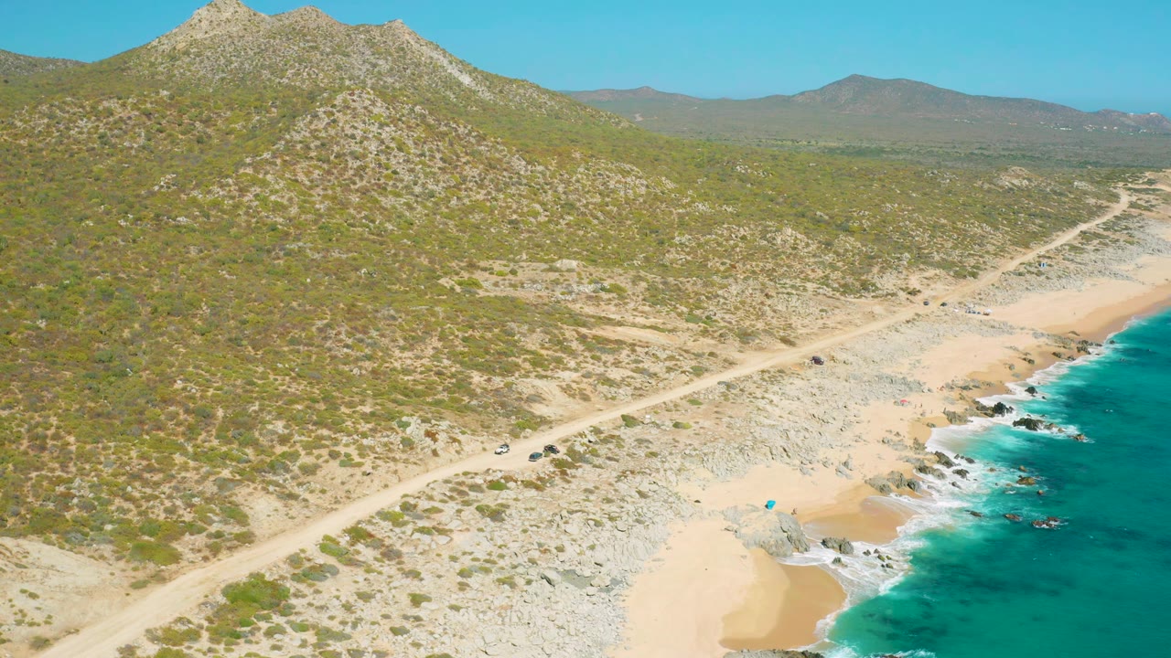 A high oblique aerial view in East Cape BCS Mexico frames the road diagonally through rocky sand, with turquoise surf along the right edge, low mountains beyond, and no visible people.