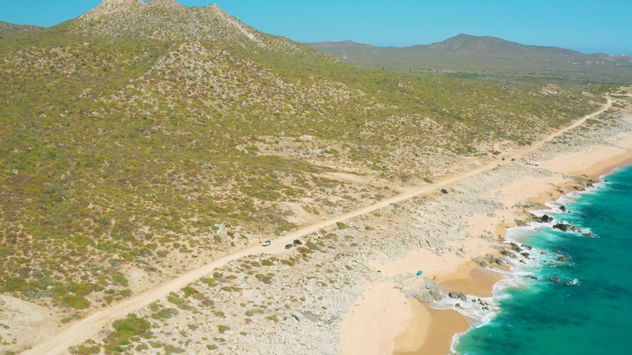The East Cape BCS Mexico coastline remains in a wide drone view, with a sandy cove and rocks below the road, bright turquoise ocean at right, inland mountains at left, and no visible people.