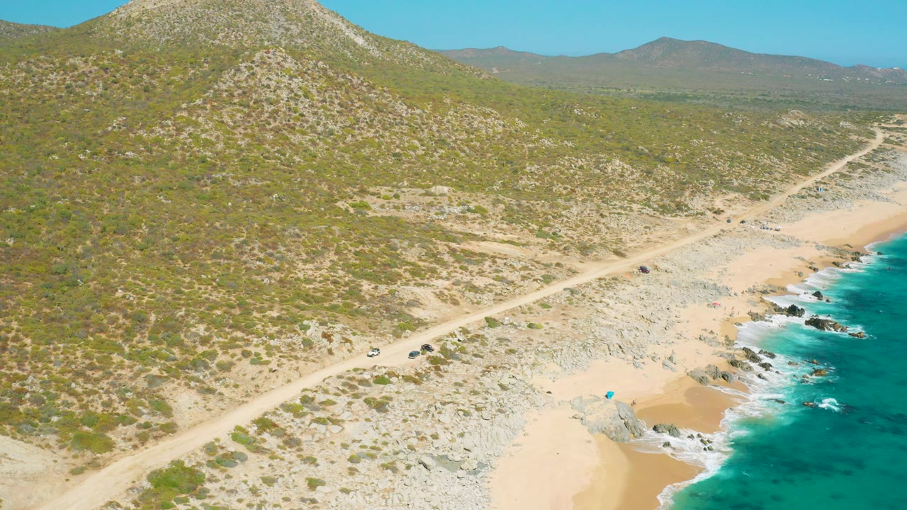 A wide aerial frame in East Cape BCS Mexico shows scrubby hillside filling the left side, the dirt road cutting across the center, rocky beach below, turquoise water at the right edge, and no visible people.