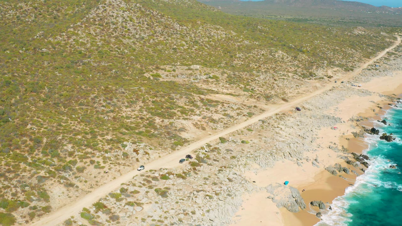 Aerial frame over East Cape, Baja California Sur, Mexico showing a pale dirt road above scrubby hills, rocky sand, turquoise surf at the right edge, a small white vehicle, and no visible people.