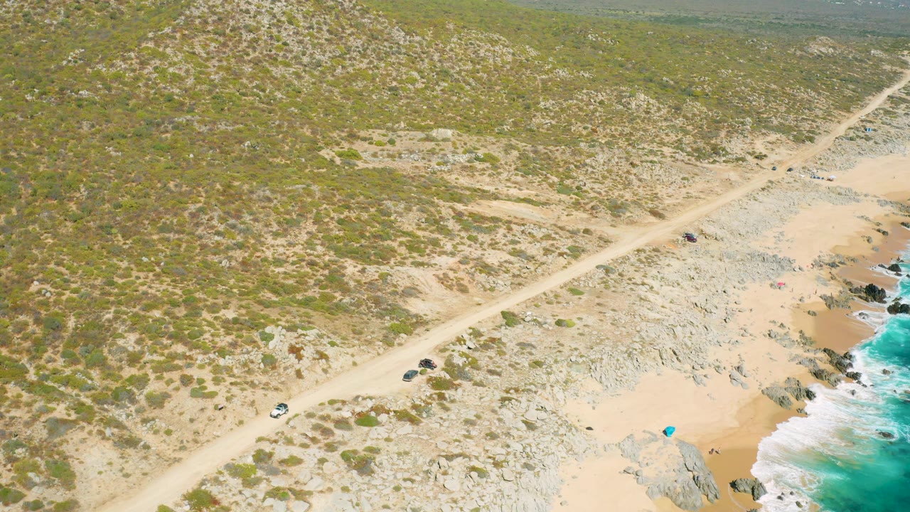 Aerial coastal road passing above a sandy beach and bright surf.