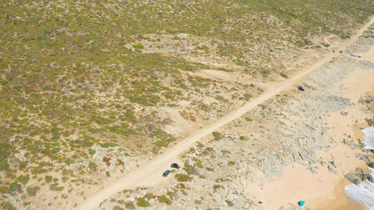 Aerial frame from East Cape BCS Mexico with the dirt road running diagonally through low green scrub, pale sand, and gray rock, with the vehicle slightly farther along the road and no visible people.