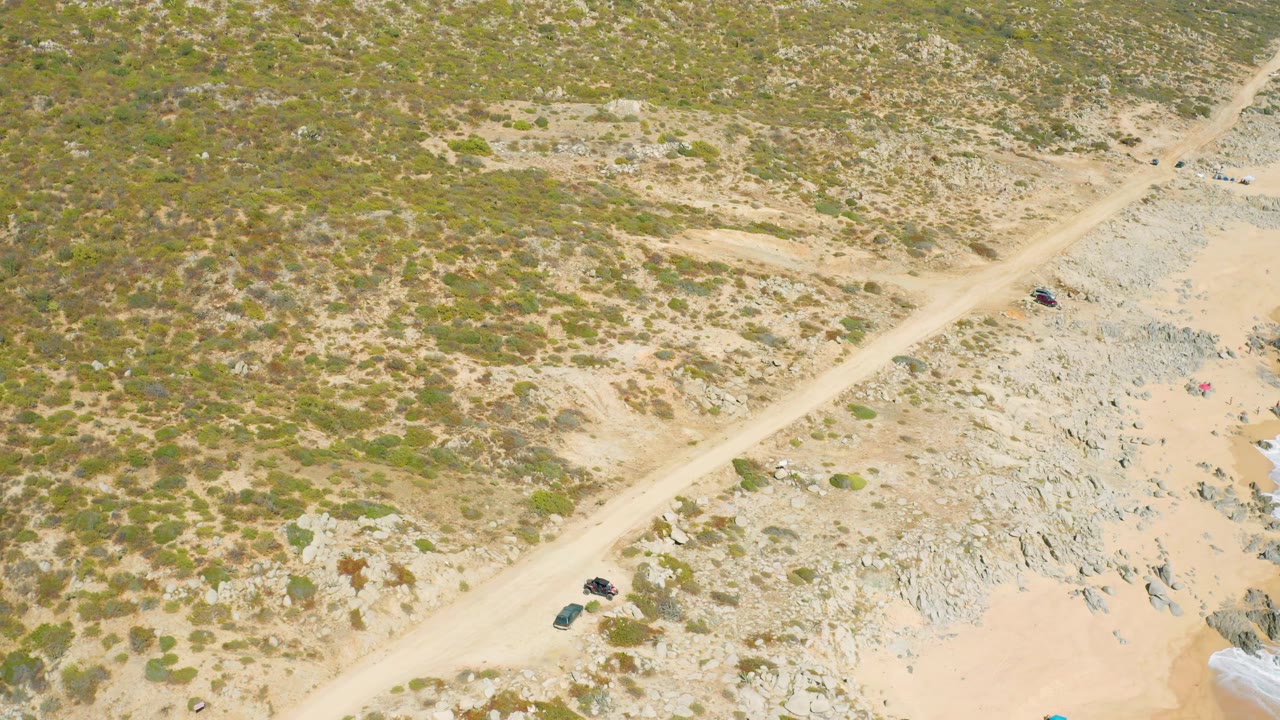Aerial frame from East Cape BCS Mexico showing the vehicle on the diagonal dirt road beside scattered scrub, rocky ground, and a small visible edge of surf, with no visible people.