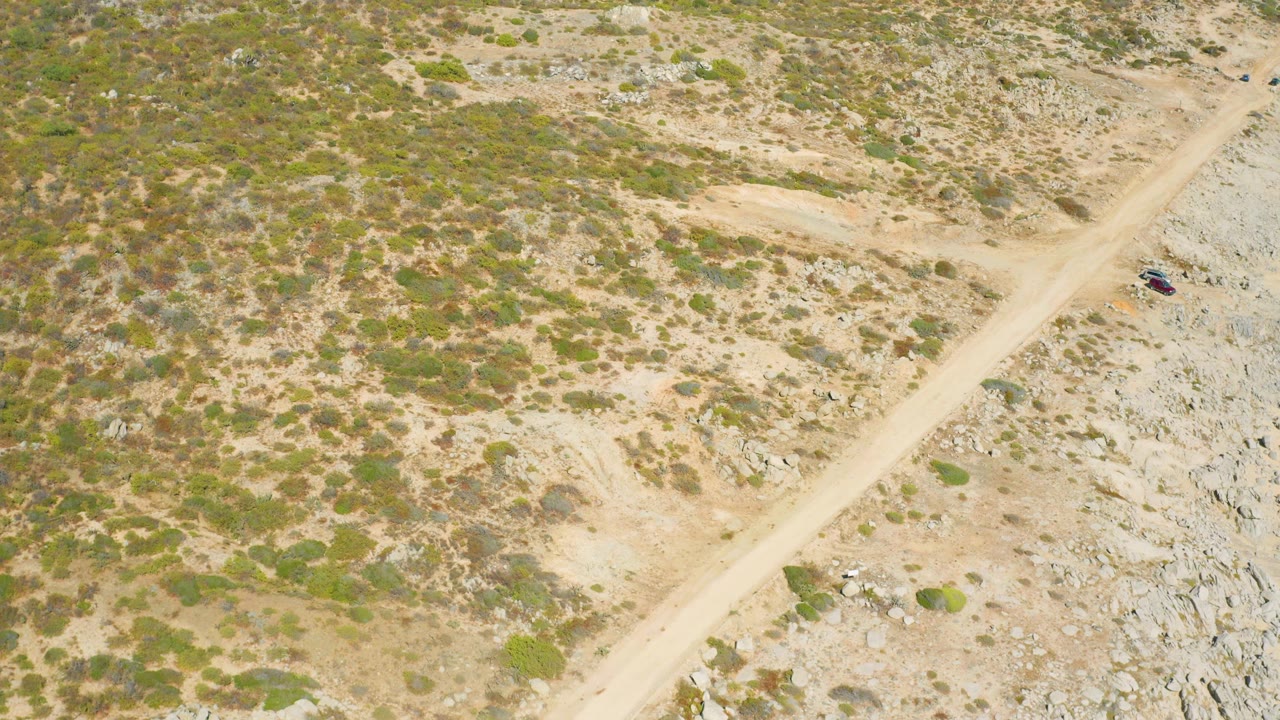 Aerial frame from East Cape BCS Mexico with the vehicle slightly farther along the dirt road, bordered by gray rocks, tan soil, and low green shrubs with no visible people.