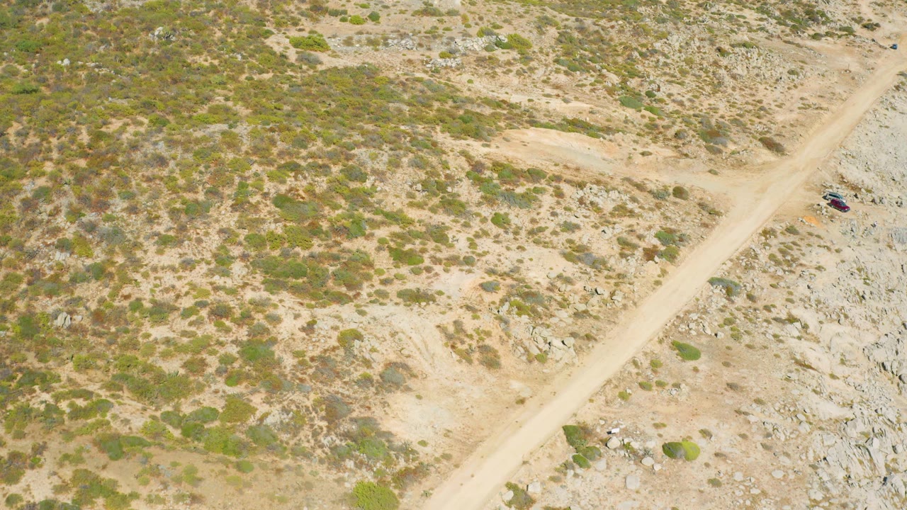 Aerial frame from East Cape BCS Mexico showing the road angled through sparse scrub, with the small vehicle close to the right edge and no visible people.