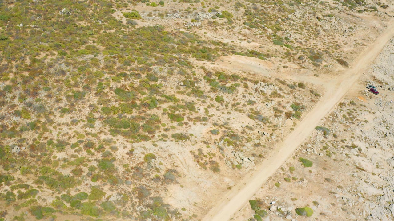 Aerial frame from East Cape BCS Mexico looking down on a pale dirt road and rocky shrubland, with the vehicle still visible on the road and no visible people.