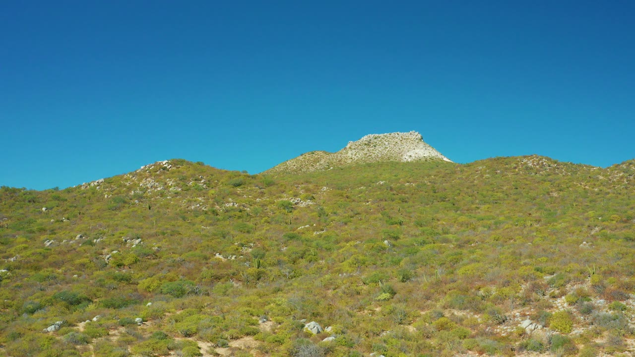 Fourth frame presents the East Cape BCS Mexico hillside from a steady wide view, with rock-strewn vegetation below a bright blue sky and no visible people.