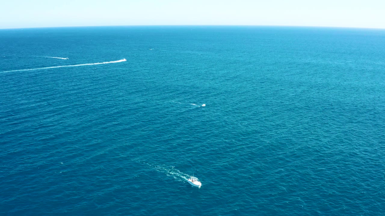Wide aerial frame of East Cape blue ocean with a small vessel and several thin white wakes spread across the water, with no visible people.