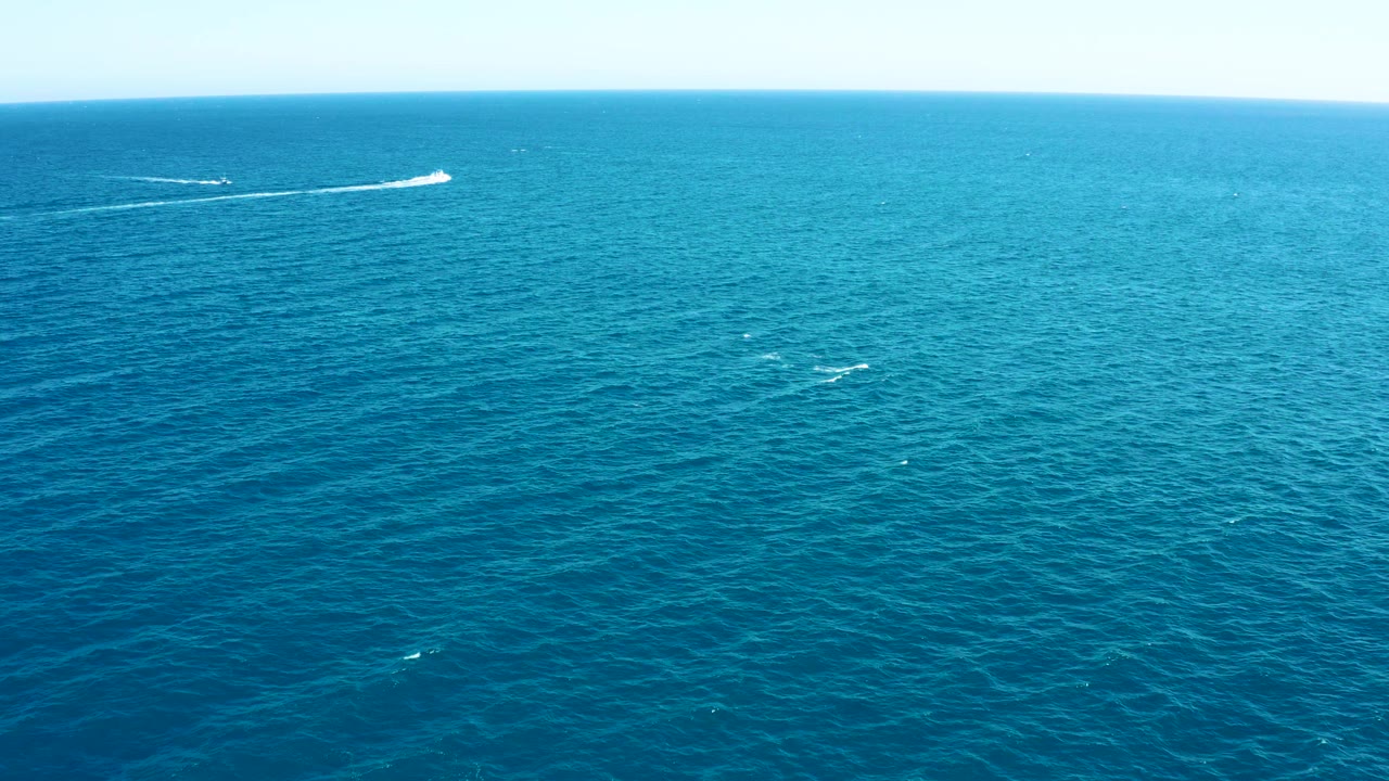 Wide aerial frame over East Cape, Baja California Sur with textured turquoise-blue ocean, a distant white boat wake near the horizon, and small surfacing marks in the midwater, with no visible people.