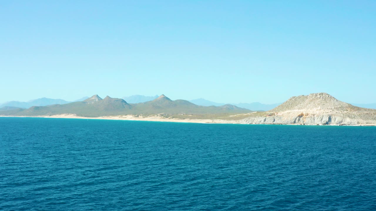 East Cape Baja California Sur ocean view with rippled blue water in front, a pale sandy beach and mountain range on the horizon, and no visible people.