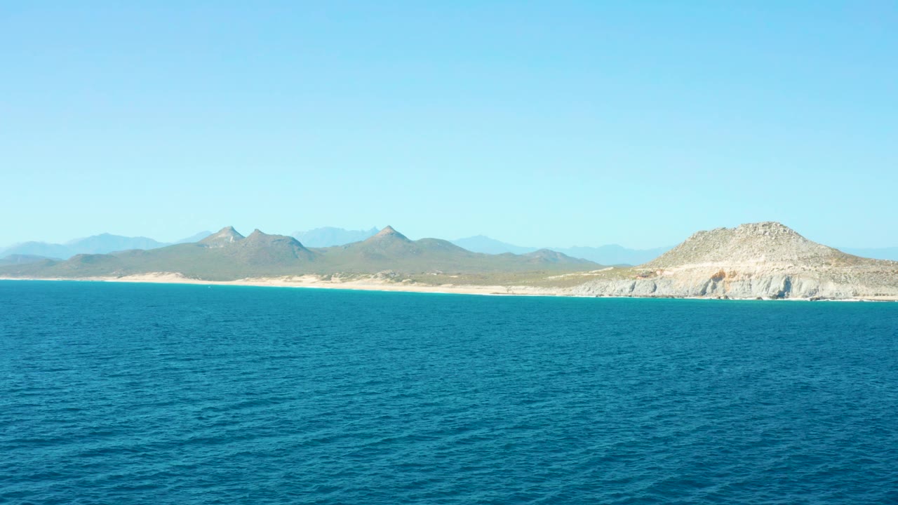 East Cape Baja California Sur coast seen across calm blue water, with the sandy shore running below a dry mountain range and no visible people.