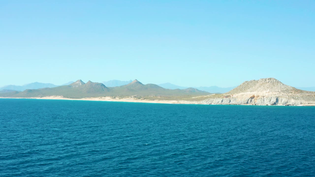 East Cape Baja California Sur shoreline appears beyond open sea, with pale sand, rocky hills, layered blue mountains, and no visible people.