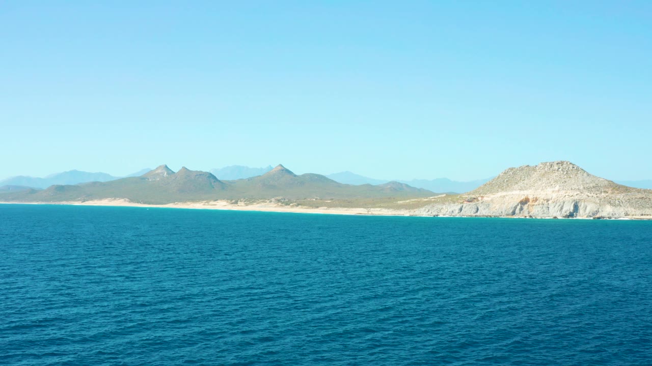 East Cape Baja California Sur water fills the foreground while the sandy coast and desert-colored mountain range stay distant under clear daylight, with no visible people.