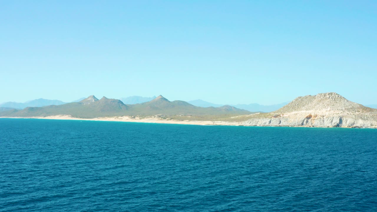 Blue ocean with sandy shoreline and distant mountains under clear sky.