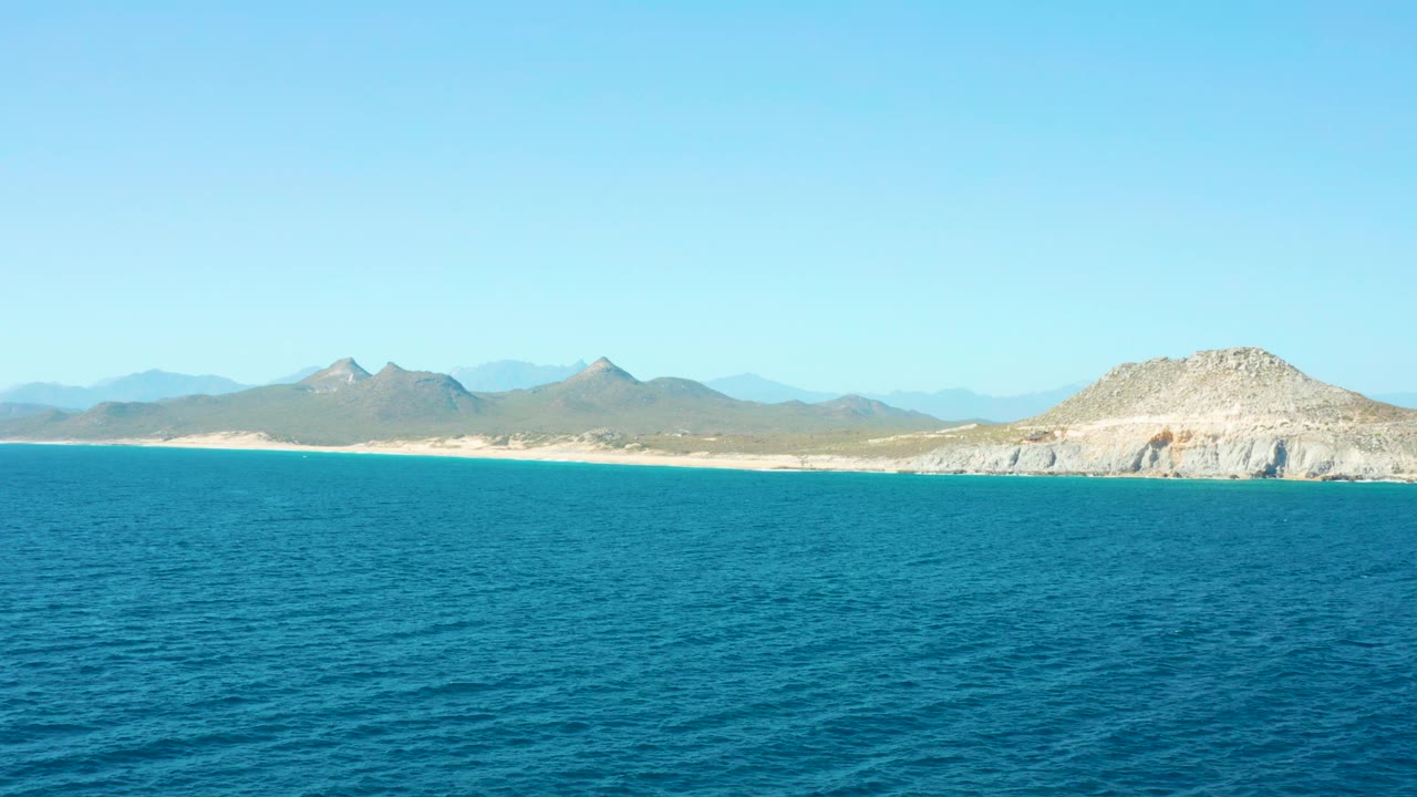 East Cape coastal view one second in, showing textured blue water, a pale beach band, low dry ridges, a larger rocky hill to the right, clear sky, and no visible people.