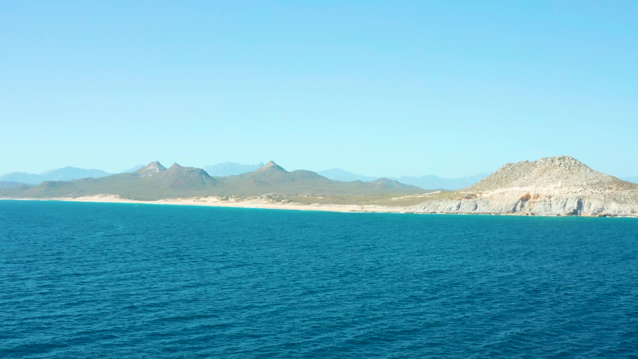 Aerial East Cape frame with open blue ocean covering the lower half, sandy coastline through the middle, layered mountains in the distance, clear daylight, and no visible people.