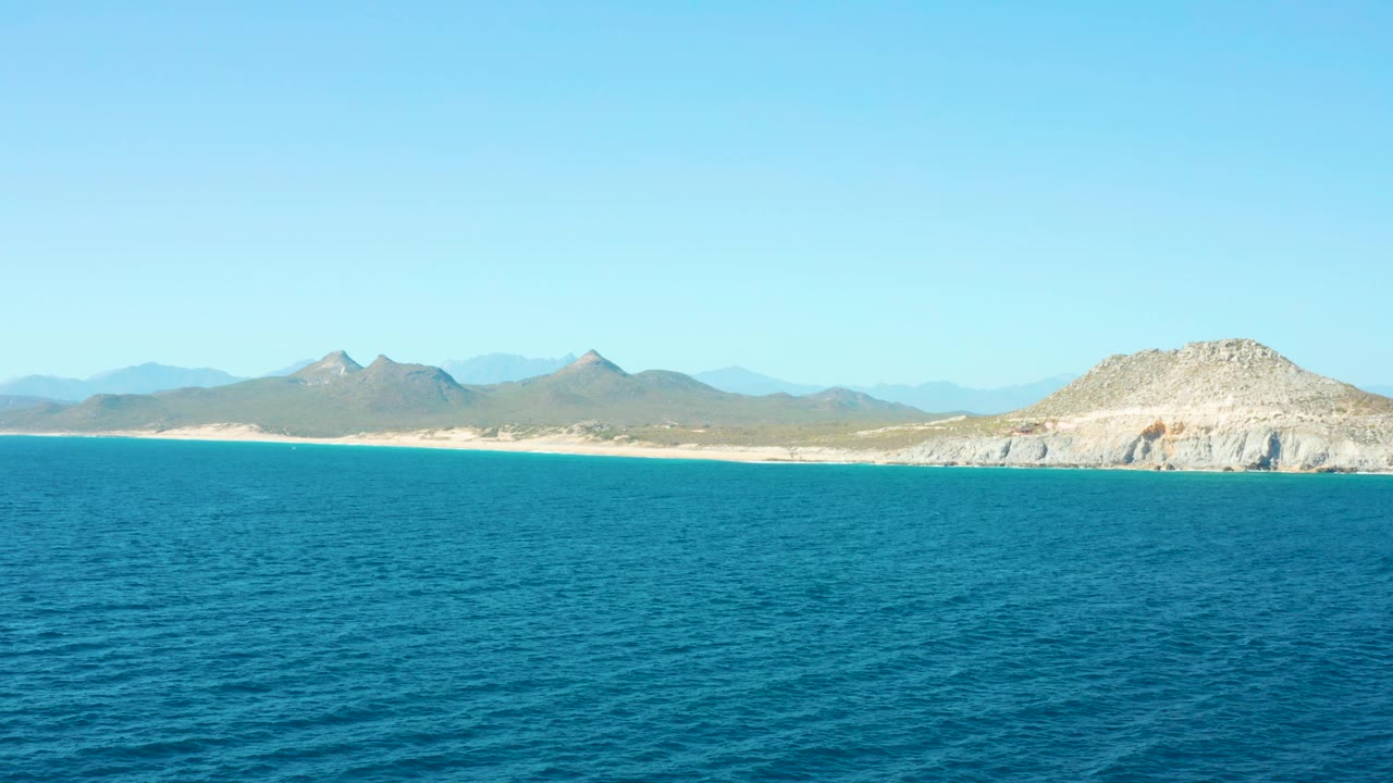 Mid-clip view of the East Cape coast, with blue sea in the foreground, a narrow sandy beach, distant dry mountains, a rocky hill near the right edge, and no visible people.