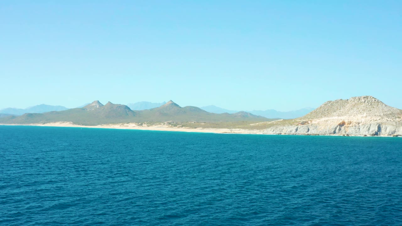 East Cape BCS Mexico shoreline one second in, showing textured blue water across the foreground, sand along the coast, distant mountains, the rocky headland at right, and no visible people.