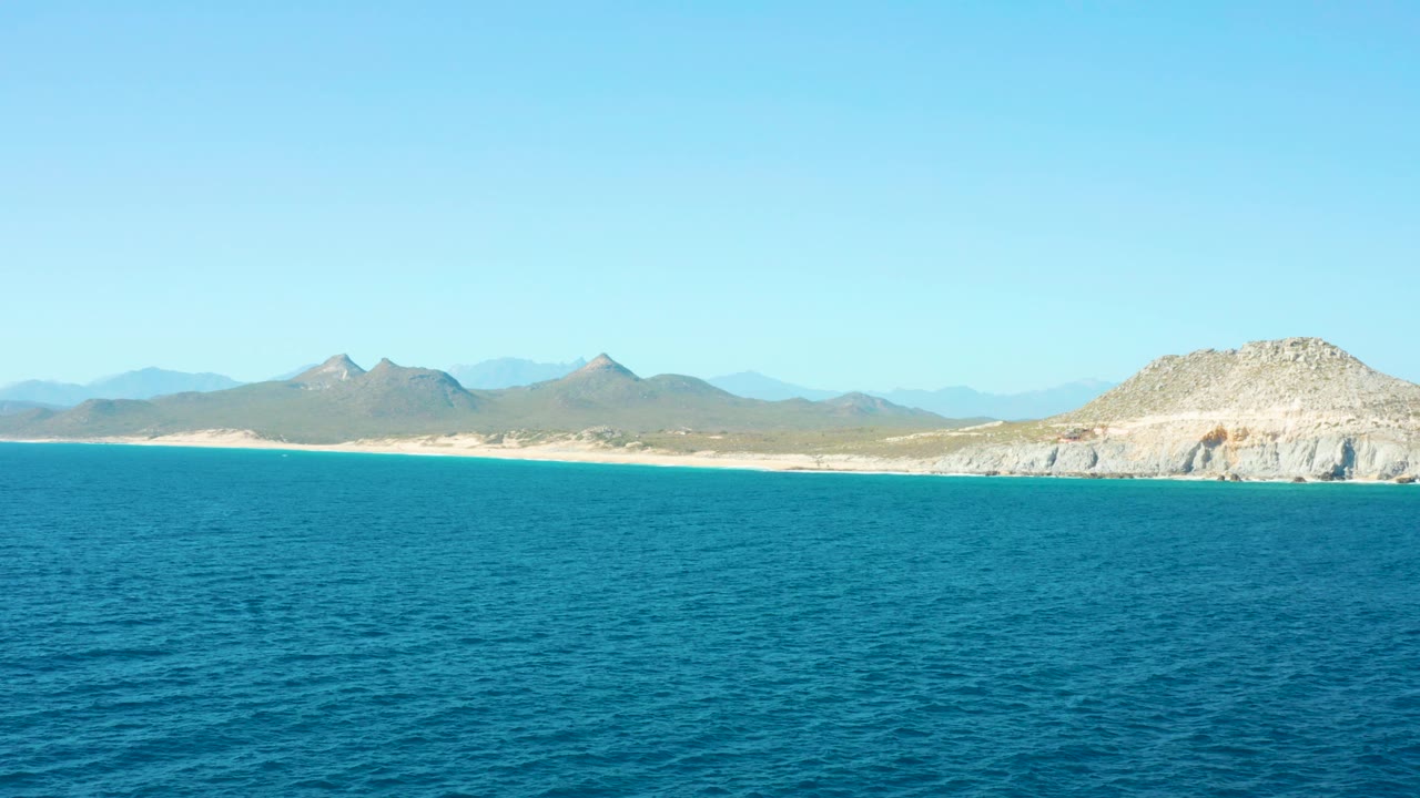 East Cape BCS Mexico wide coastal frame with the ocean occupying most of the lower image, a thin sandy beach at the horizon, mountains behind it, a pale rocky outcrop to the right, and no visible people.