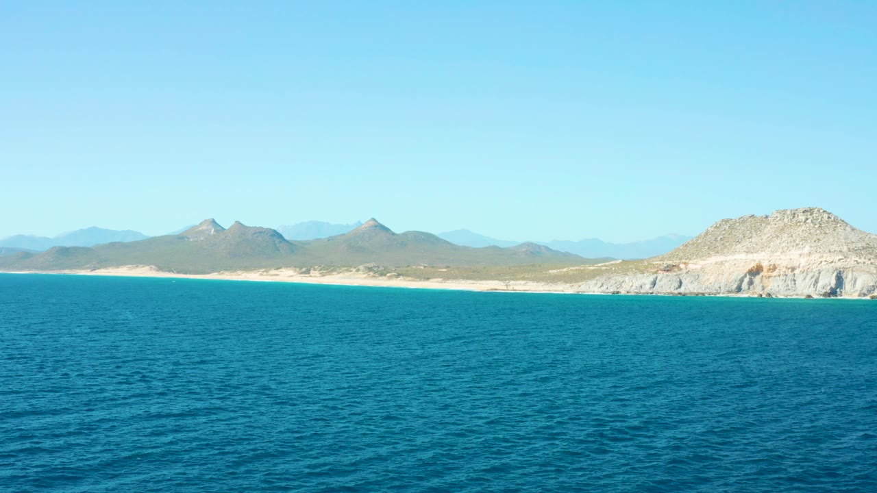Ocean surface leading to a sandy beach and layered mountains under clear sky.