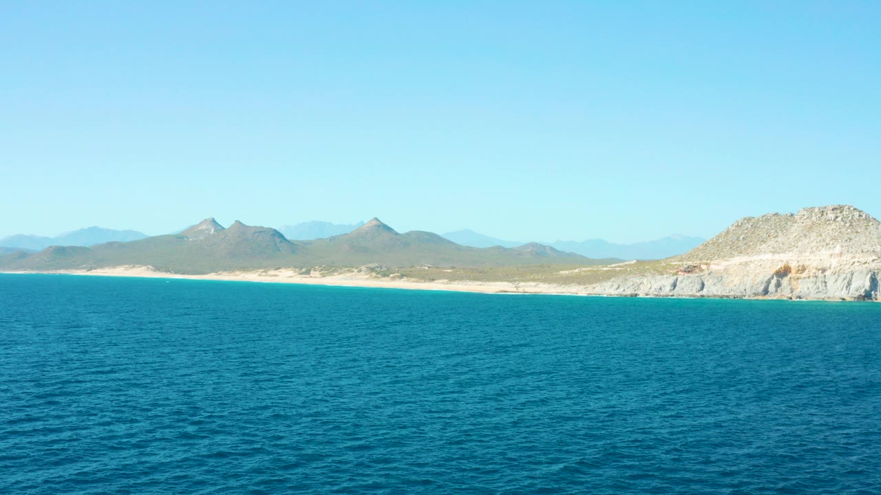 East Cape BCS Mexico coastline with blue ocean in the foreground, a sandy beach and low mountains beyond, rocky cliffs on the right, and no visible people.
