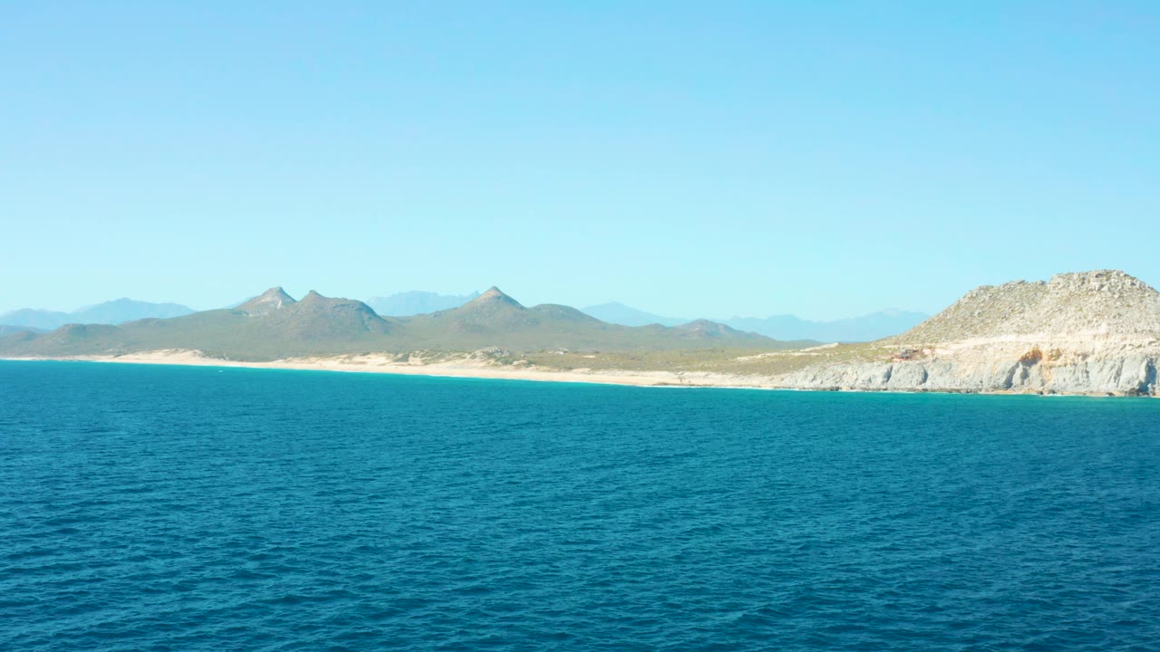 Calm blue water with sandy shoreline, mountains, and right-side cliff