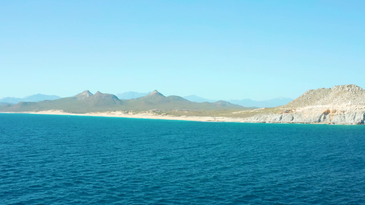 East Cape BCS Mexico shoreline seen across a wide blue ocean foreground, with a narrow sandy beach, layered mountains, pale rocky cliff on the right, and no visible people.