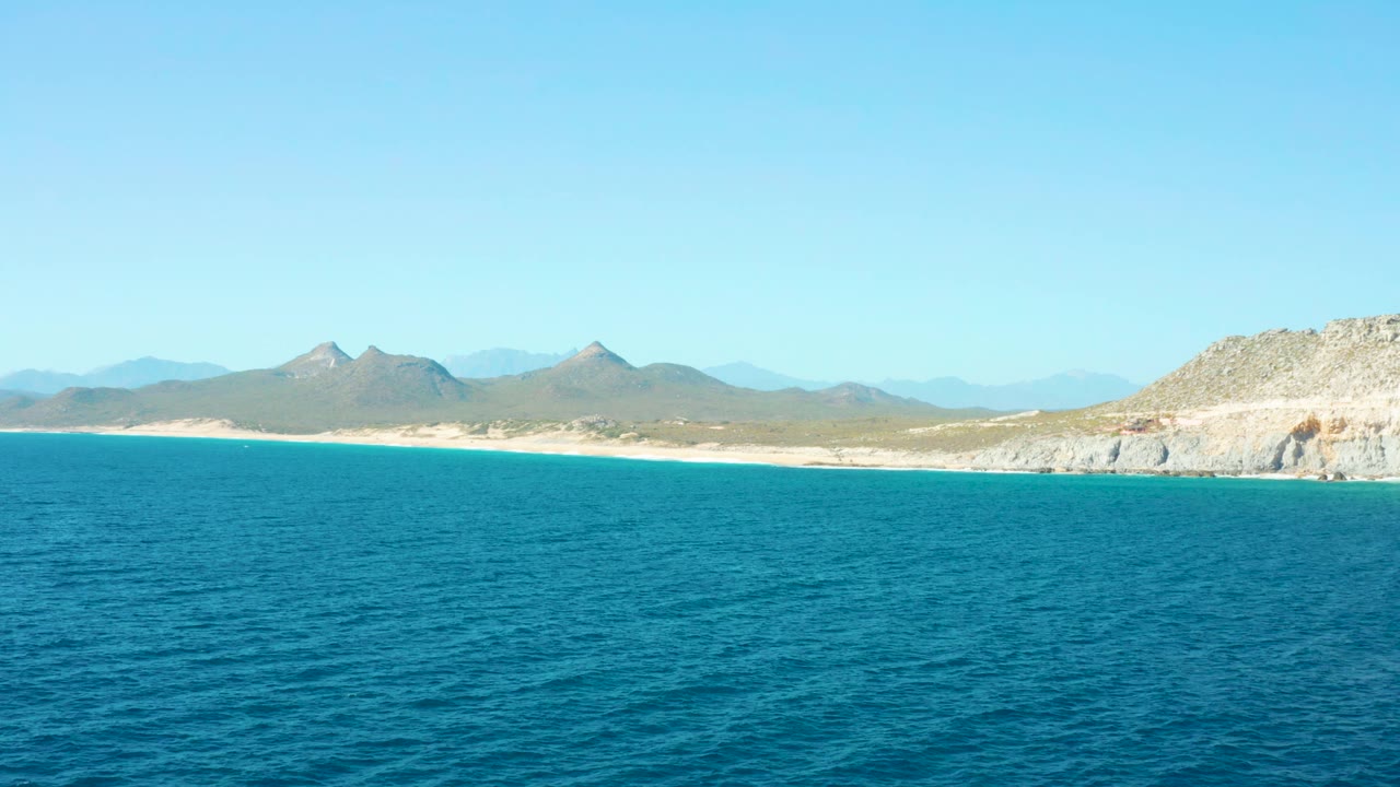 East Cape BCS Mexico coastline remains wide in frame, with textured blue water below, a narrow sand strip, rocky land at right, layered mountains, and no visible people.