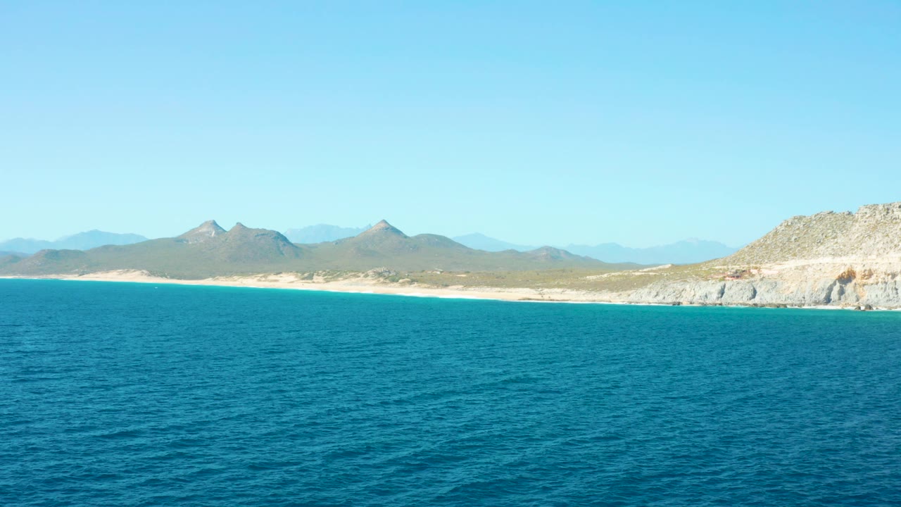 Aerial coastal frame from East Cape BCS Mexico showing rippled blue water, pale beach, scrubby land, low mountains across the horizon, clear blue sky, and no visible people.