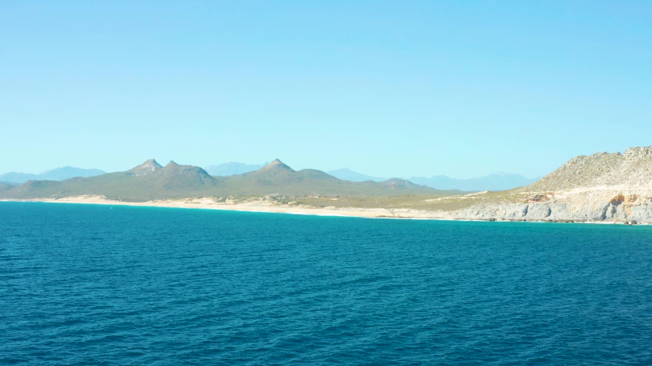 East Cape BCS Mexico beach and mountains sit beyond a large blue ocean foreground, with the rocky coastline still visible at right and no visible people in the frame.