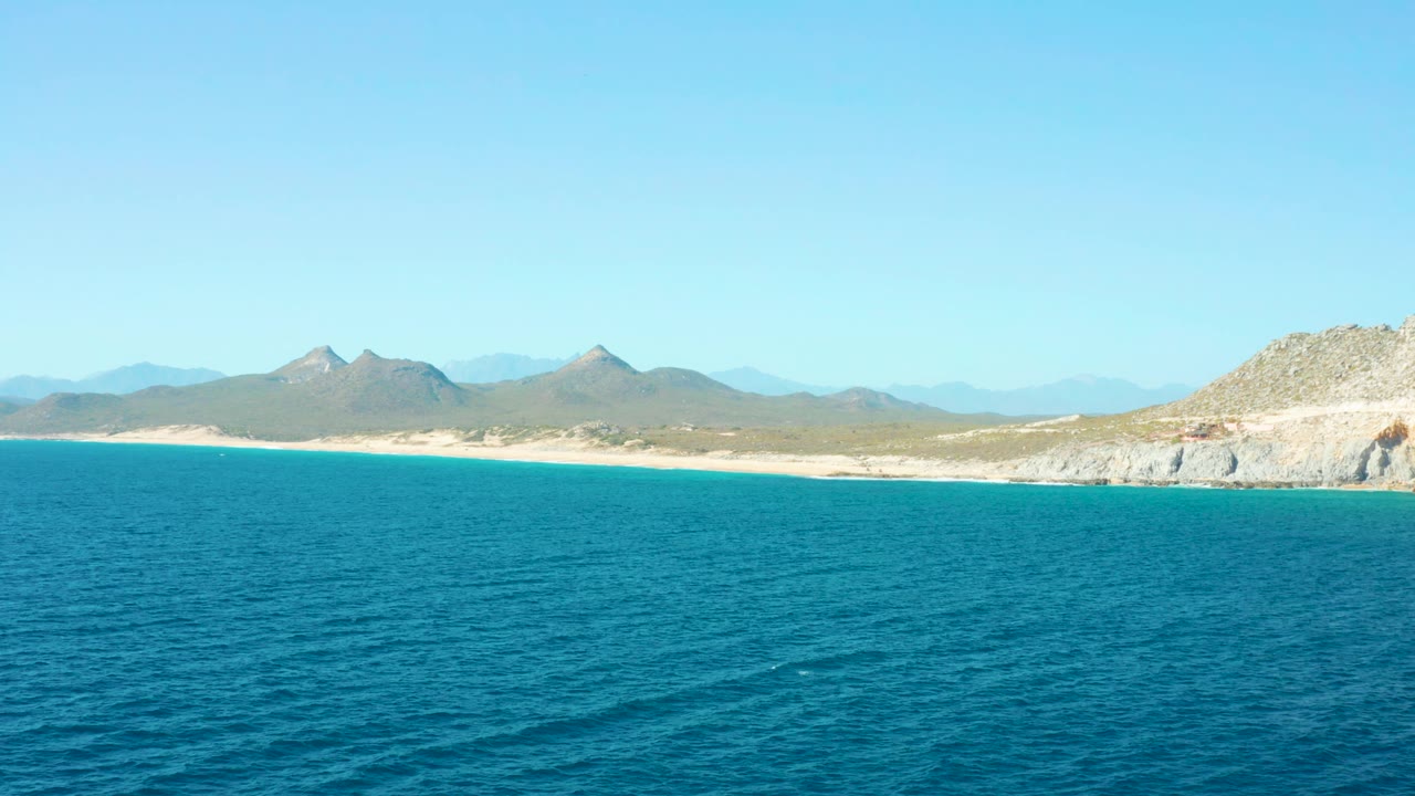 East Cape ocean surface fills the lower frame while a pale sandy shoreline and layered Baja California Sur mountains sit under a clear sky with no visible people.